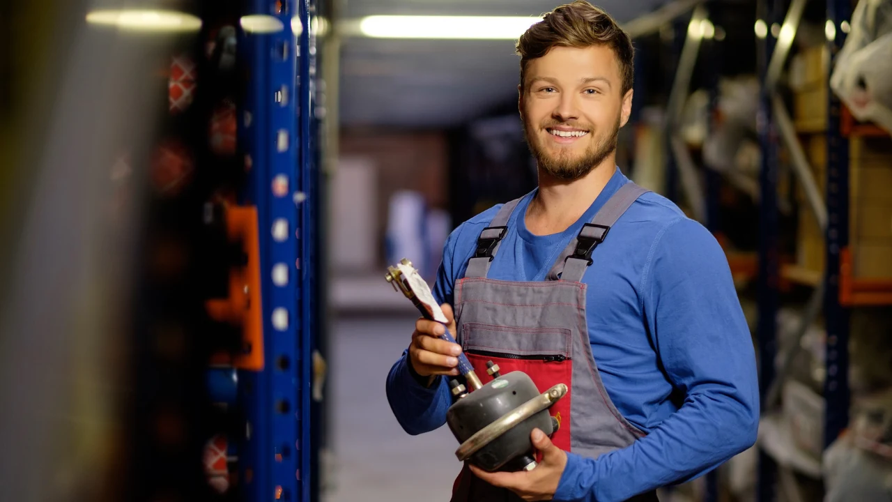 Maintenance technician in a parts storeroom holding a spare brake chamber pulled from the shelves
