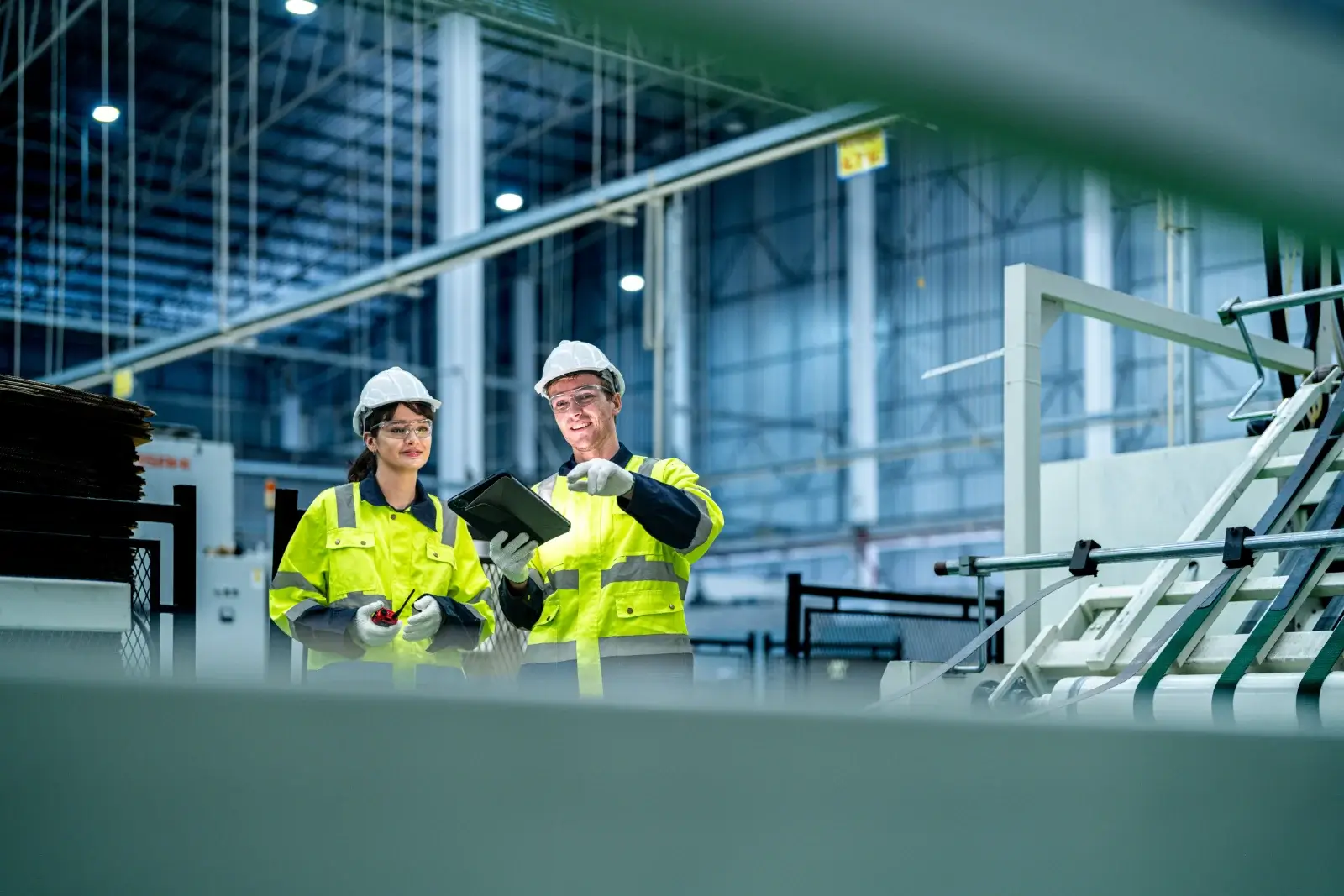 Two plant technicians in hard hats reviewing maintenance records on a tablet on a manufacturing floor