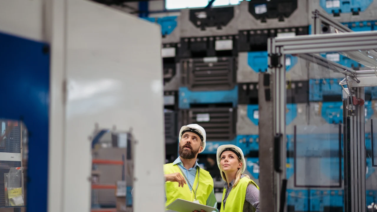 Two technicians in hard hats on a warehouse floor checking equipment with a tablet in hand